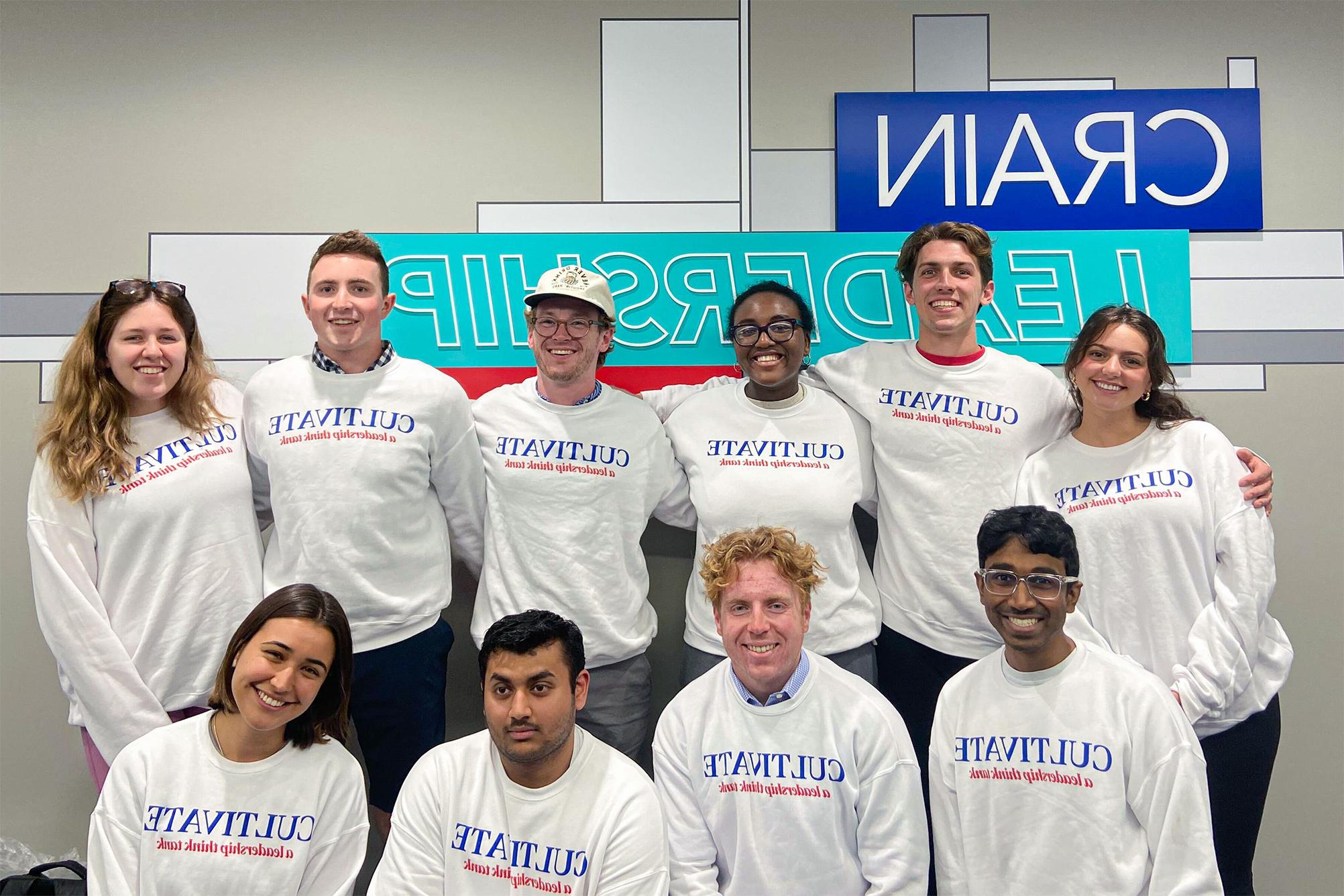 group of students in white sweatshirt with "Cultivate" in blue ink and "a leadership think tank" in red ink, standing in front gray wall of sign "Crain Leadership"