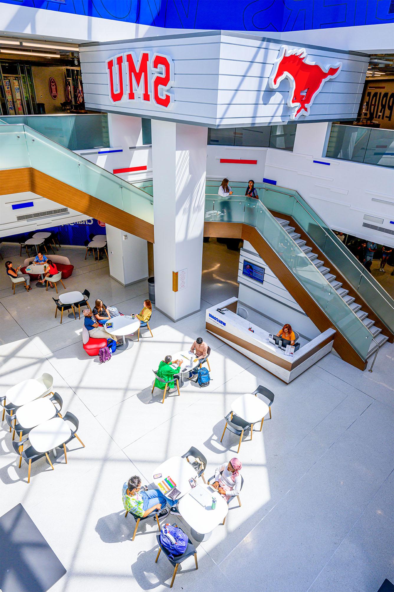 indoor atrium of building with students sitting at tables, a large staircase to the next floor, a red mustang cutout and SMU in red hanging on wall